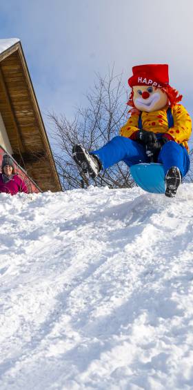 Kinder und Familotel Maskottchen Happy rodeln fröhlich im verschneiten Familienhotel Mein Krug