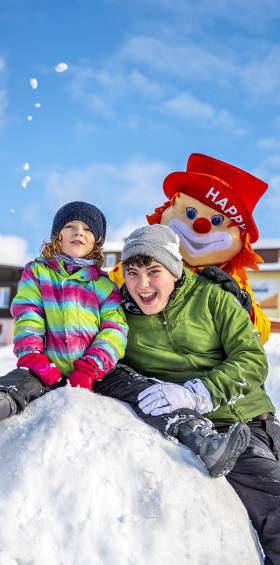 Familie mit dem Familotel Maskottchen Happy auf einem großen Schneeball vor dem Hotel Mein Krug