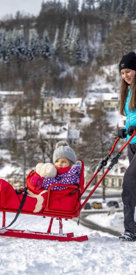 Mutter zieht ein Kleinkind in einem roten Schlitten durch eine verschneite Winterlandschaft