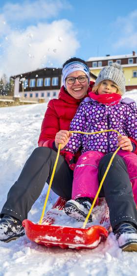 Familie genießt eine Schlittenfahrt auf einer schneebedeckten Abfahrt vor dem Hotel Mein Krug