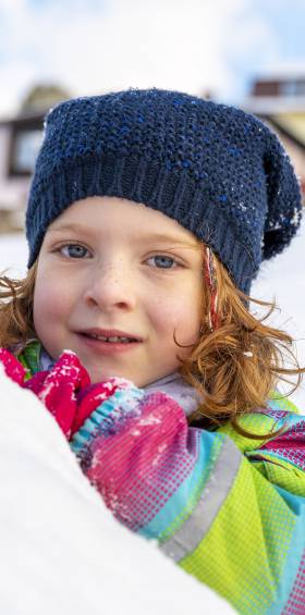 Kleines Mädchen in Winterkleidung schaut vergnügt hinter einem Schneehügel hervor.